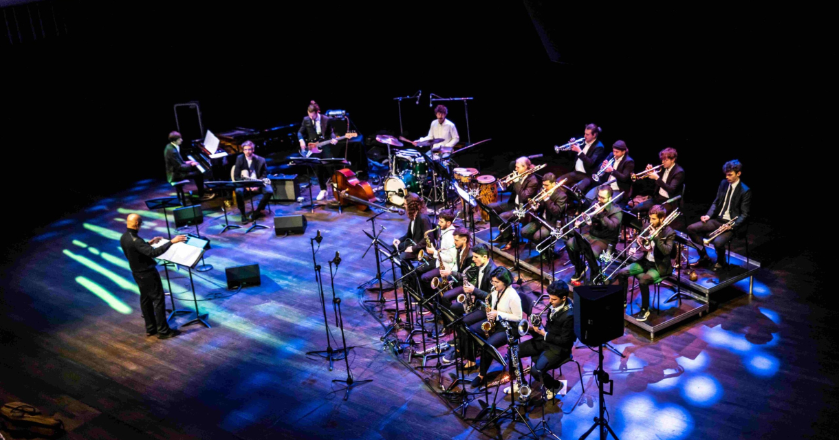 De KC Big Band treedt op onder leiding van een dirigent, met een volledig ensemble van blazers, ritmesectie en percussie. Kleurrijke verlichting en een donkere zaal versterken de dynamiek van dit jazzoptreden op het podium De KC Big Band treedt op onder leiding van een dirigent, met een volledig ensemble van blazers, ritmesectie en percussie. Kleurrijke verlichting en een donkere zaal versterken de dynamiek van dit jazzoptreden op het podium