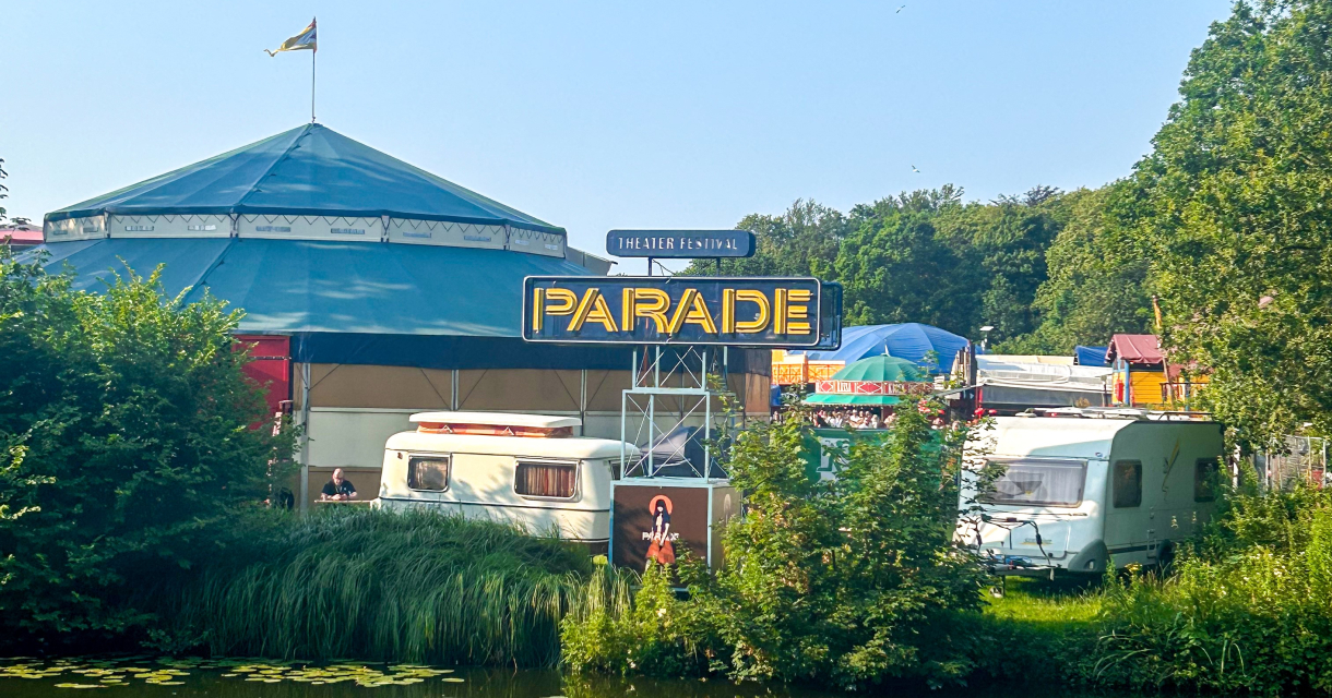 Circustenten en kleurrijke wagens van festival De Parade staan opgesteld in een groen parklandschap, weerspiegeld in het rustige water van een vijver onder een heldere blauwe lucht. Circustenten en kleurrijke wagens van festival De Parade staan opgesteld in een groen parklandschap, weerspiegeld in het rustige water van een vijver onder een heldere blauwe lucht.