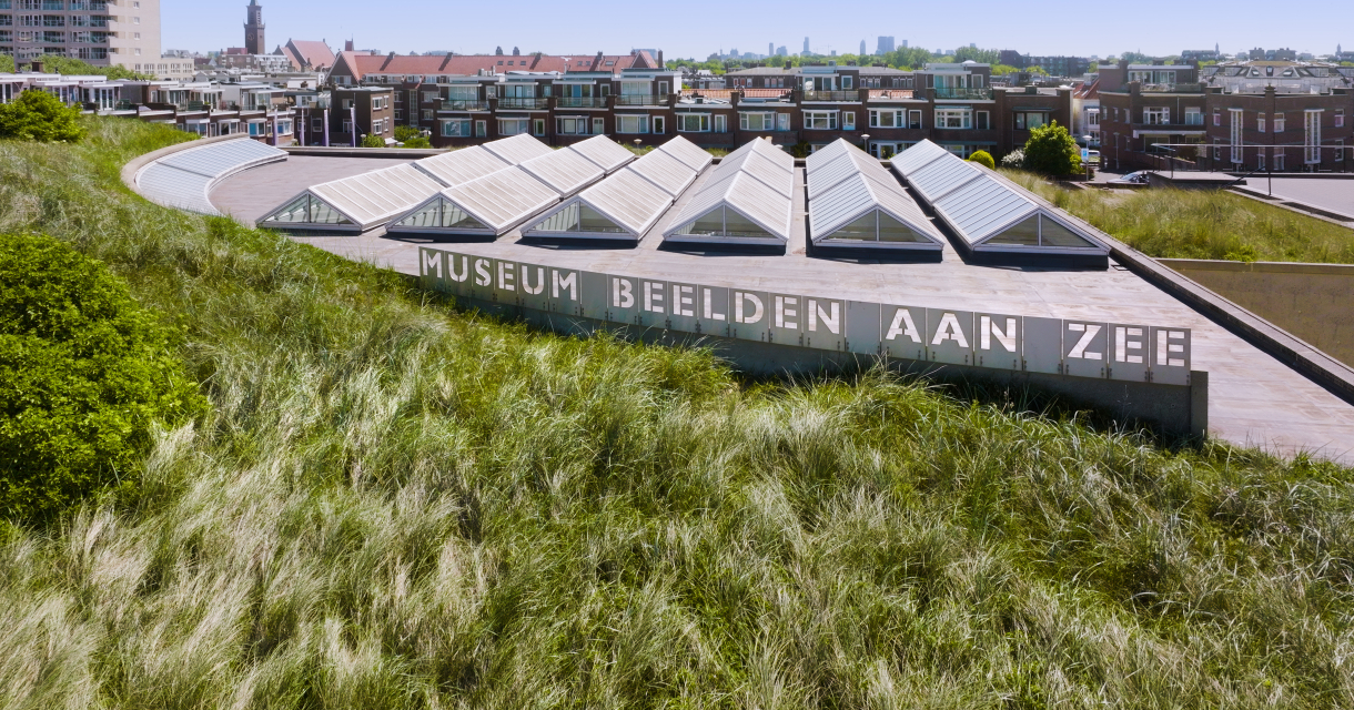 Luchtfoto van Museum Beelden aan Zee in Scheveningen, met een grasduin op het dak en de skyline van Den Haag op de achtergrond.