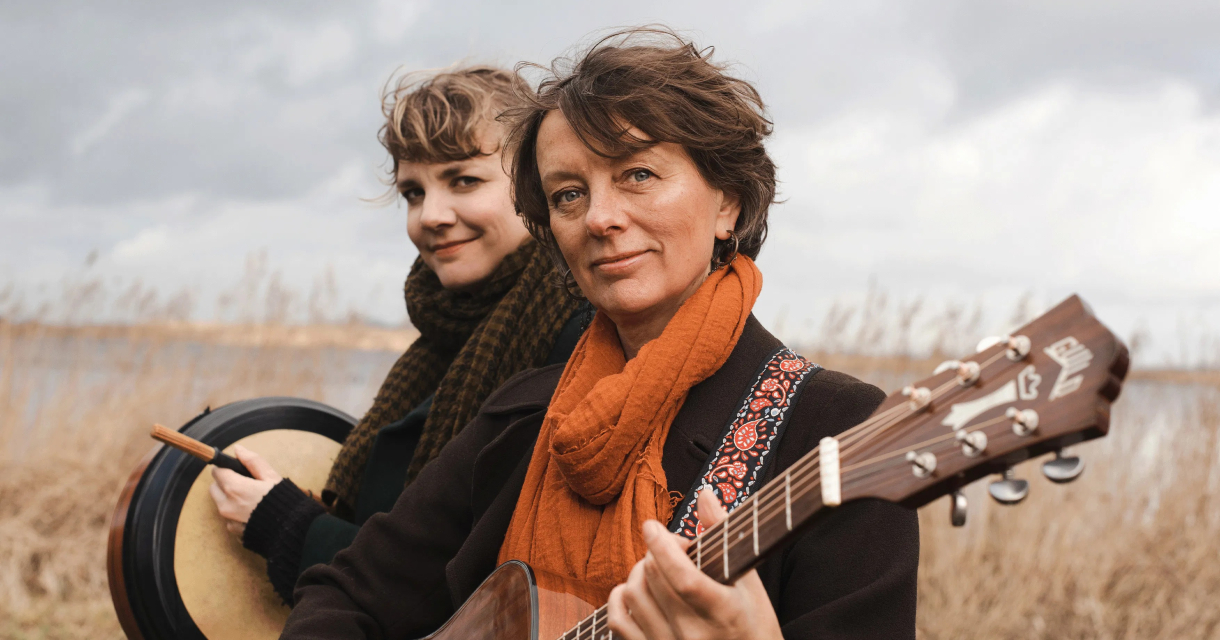 Twee muzikanten buiten bij riet en water, één met gitaar en oranje sjaal, de ander met een trommel en stok. Twee muzikanten buiten bij riet en water, één met gitaar en oranje sjaal, de ander met een trommel en stok.