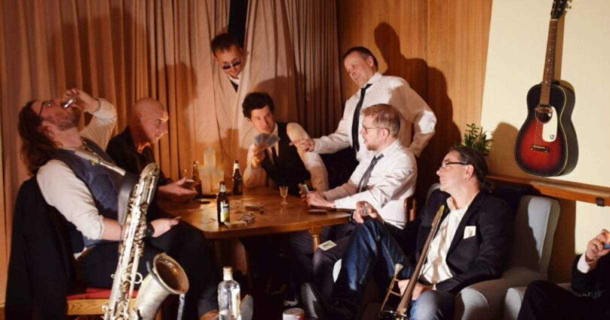 De band in een setting vol rock-’n-roll en jaren 20-swing met kaartspel, drankje en gitaar aan de muur. De band in een setting vol rock-’n-roll en jaren 20-swing met kaartspel, drankje en gitaar aan de muur.
