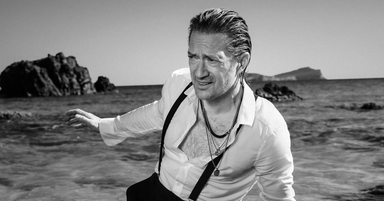 Zwart wit foto van Joost Spijkers die uit de zee komt lopen. Hij staat op een tropisch strand met rotsen en draagt een zwarte broek met bretels en een witte blouse. Promotiebeeld voor zijn show: De broer van Aznavour. Zwart wit foto van Joost Spijkers die uit de zee komt lopen. Hij staat op een tropisch strand met rotsen en draagt een zwarte broek met bretels en een witte blouse. Promotiebeeld voor zijn show: De broer van Aznavour.