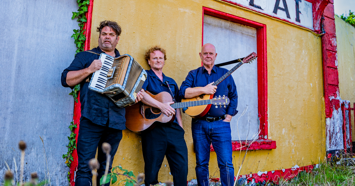 Drie mannen met accordeon, gitaar en bouzouki poseren voor een vervallen geel-rood gebouw met het woord "CAF" op de gevel. Drie mannen met accordeon, gitaar en bouzouki poseren voor een vervallen geel-rood gebouw met het woord "CAF" op de gevel.