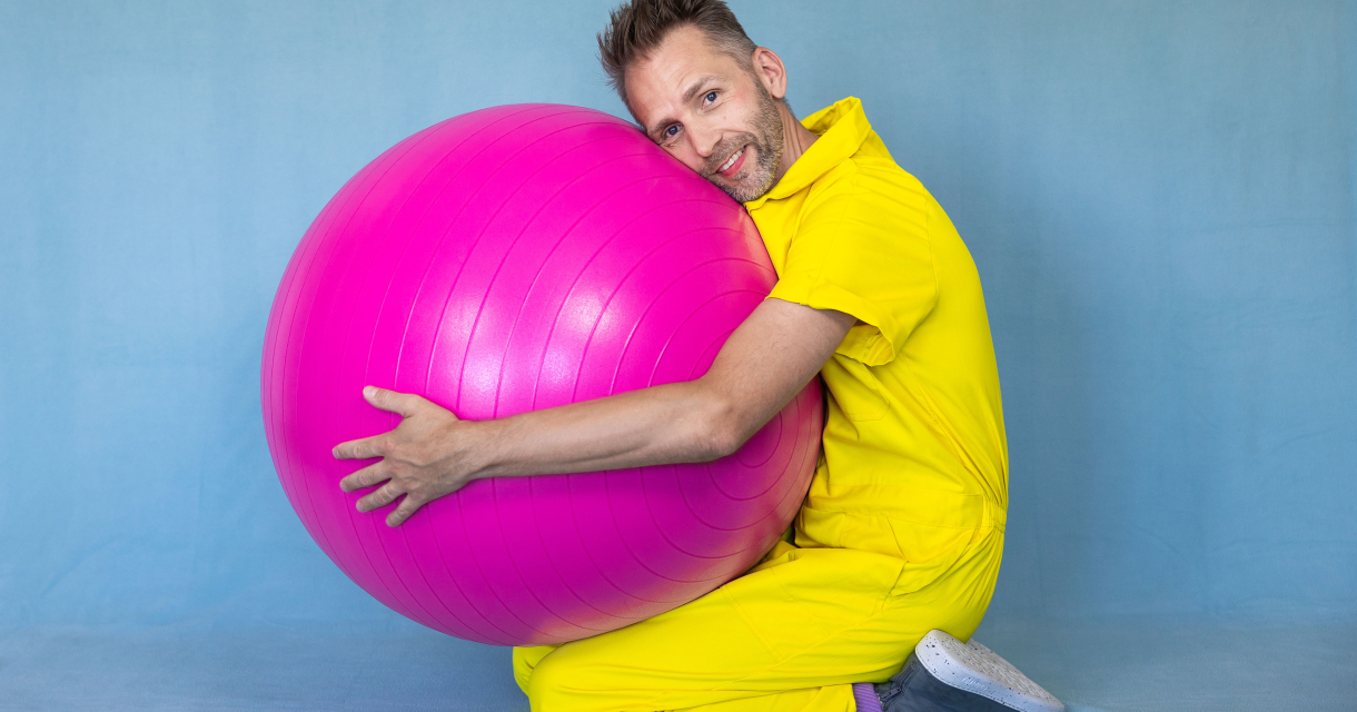 Promotiebeeld voor Peutervoorstelling Rokkerol Efrem Stein zit op de grond in een gele overal en knuffelt een grote roze bal Promotiebeeld voor Peutervoorstelling Rokkerol Efrem Stein zit op de grond in een gele overal en knuffelt een grote roze bal