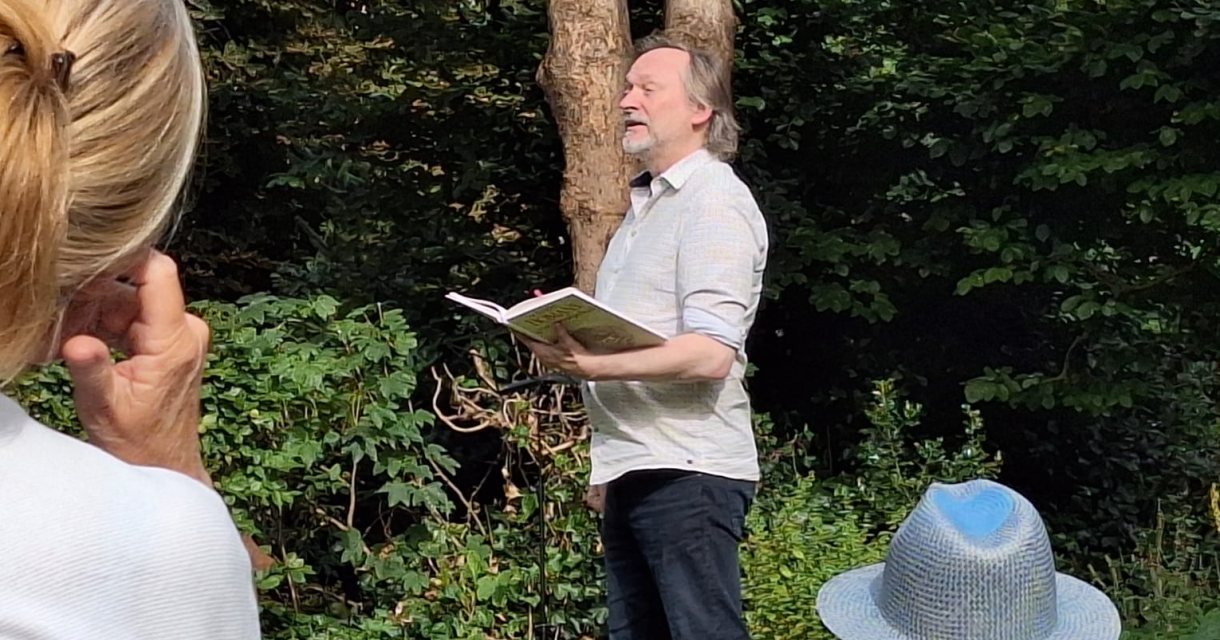 Man leest voor uit een boek tijdens een buitenoptreden in een groene, bosrijke omgeving; toeschouwers luisteren vanaf stoelen op het gras. Man leest voor uit een boek tijdens een buitenoptreden in een groene, bosrijke omgeving; toeschouwers luisteren vanaf stoelen op het gras.