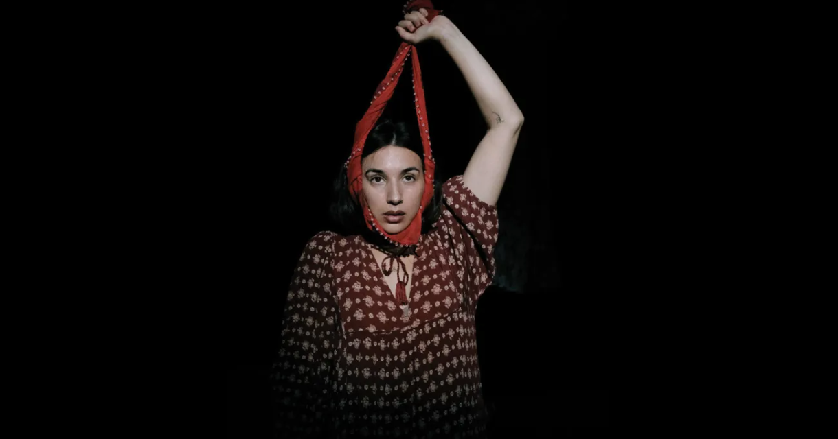 Promotiebeeld dansclick Melisa Diktaş in een rode bloemenjurk kijkt recht in de camera, met een rode stoffen band strak rond haar gezicht en omhooggehouden arm, tegen een donkere achtergrond. Promotiebeeld dansclick Melisa Diktaş in een rode bloemenjurk kijkt recht in de camera, met een rode stoffen band strak rond haar gezicht en omhooggehouden arm, tegen een donkere achtergrond.