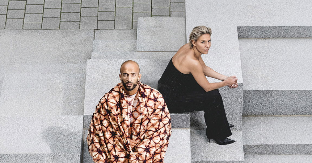 Foto van Mouna en Walid van schuin bovenaf. Ze zitten op grijze betonnen blokken en kijken recht in de camera. Mouna draagt een zwarte jumpsuit en khalid een rood-oranje tie-dye jas. Foto van Mouna en Walid van schuin bovenaf. Ze zitten op grijze betonnen blokken en kijken recht in de camera. Mouna draagt een zwarte jumpsuit en khalid een rood-oranje tie-dye jas.