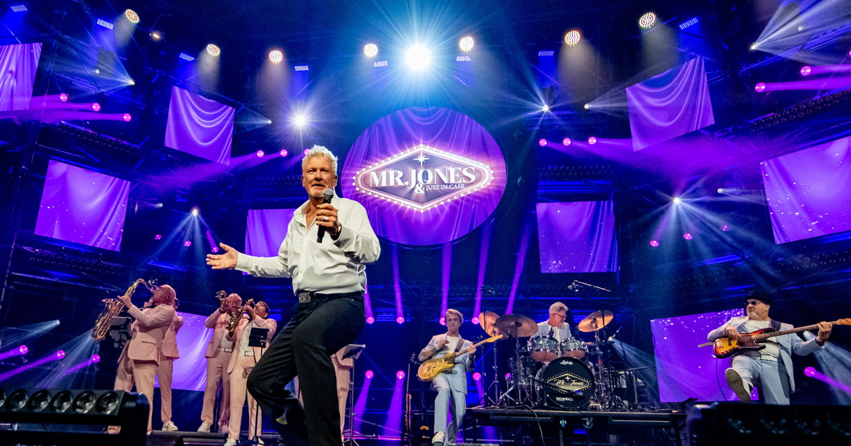 De leden van de tributeband Mr. Jones en Just in case staan op een paars verlicht podium. De leden van de tributeband Mr. Jones en Just in case staan op een paars verlicht podium.
