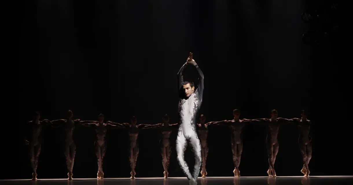 Danser in zilverkleurig kostuum op een donker podium, met rij dansers in bruine outfits op de achtergrond Danser in zilverkleurig kostuum op een donker podium, met rij dansers in bruine outfits op de achtergrond