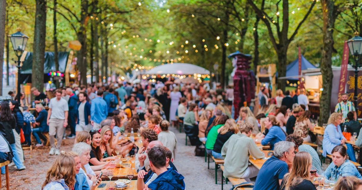 Drukke markt met lange picknicktafels onder bomen, mensen eten en drinken onder sfeerverlichting Drukke markt met lange picknicktafels onder bomen, mensen eten en drinken onder sfeerverlichting