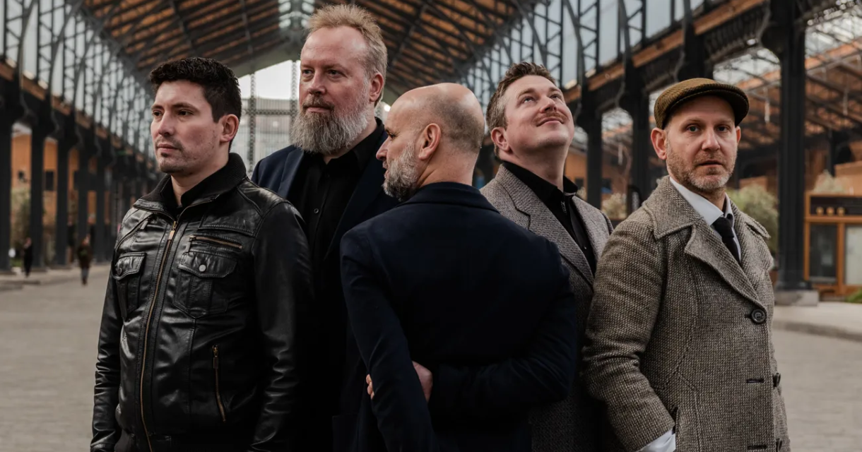 De leden van sonico, de rovira tributeband, staan in grijze en zwarte outfits in de hal van een treinstation.  
