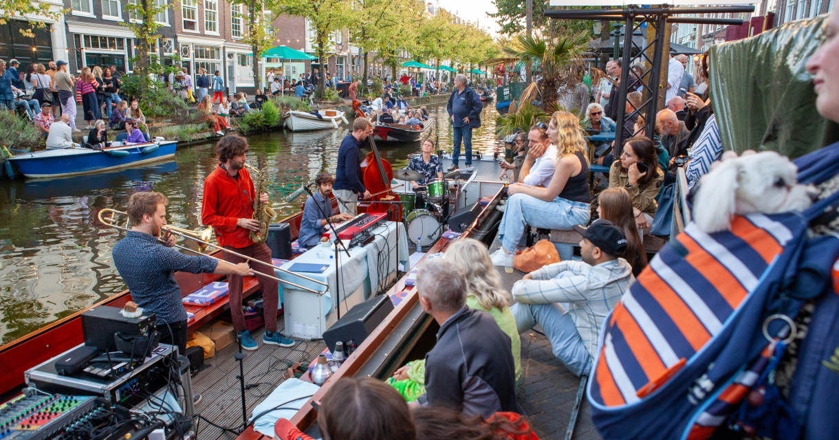Muzikanten spelen op een boot in een gracht, publiek zit langs de kade bij het Heineken Stage-bord Muzikanten spelen op een boot in een gracht, publiek zit langs de kade bij het Heineken Stage-bord