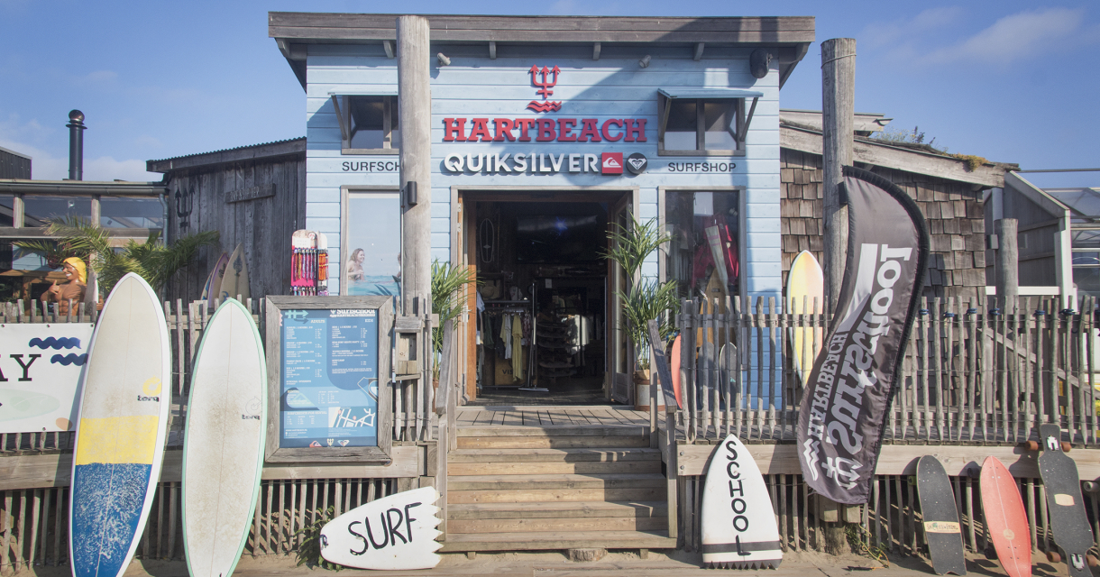 Ingang Hart Beach surfschool bestaande uit lichtblauwe houten panelen en rood logo. Er staan surfplanken op een rij voor de deur naast de trap. Ingang Hart Beach surfschool bestaande uit lichtblauwe houten panelen en rood logo. Er staan surfplanken op een rij voor de deur naast de trap.