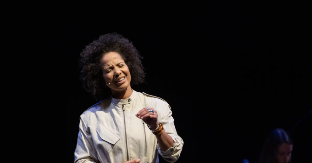 Vrouw in wit kostuum legt emotioneel iets uit op donker podium