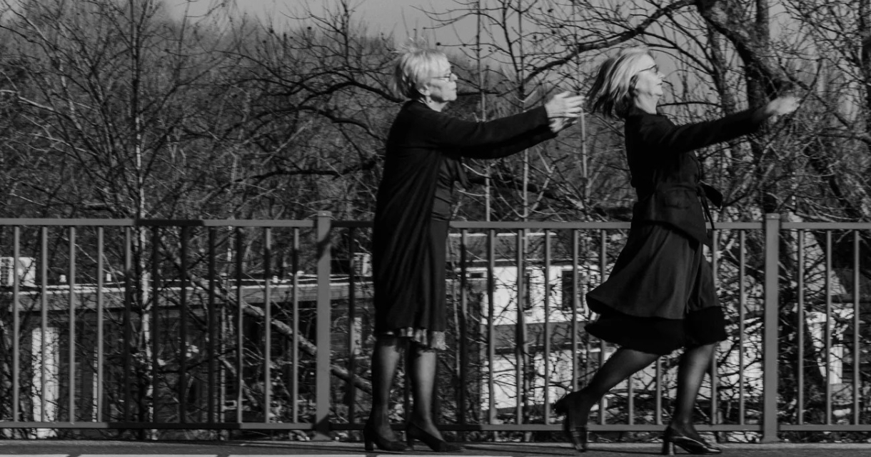 Zwart-wit promotiebeeld voorstelling dubbelspoor: Twee oudere vrouwen met witgrijs haar bevinden zich op het perron van een treinstation. Een van de vrouwen glipt weg terwijl de andere vrouw haar armen naar haar toereikt om haar te vangen. Zwart-wit promotiebeeld voorstelling dubbelspoor: Twee oudere vrouwen met witgrijs haar bevinden zich op het perron van een treinstation. Een van de vrouwen glipt weg terwijl de andere vrouw haar armen naar haar toereikt om haar te vangen.