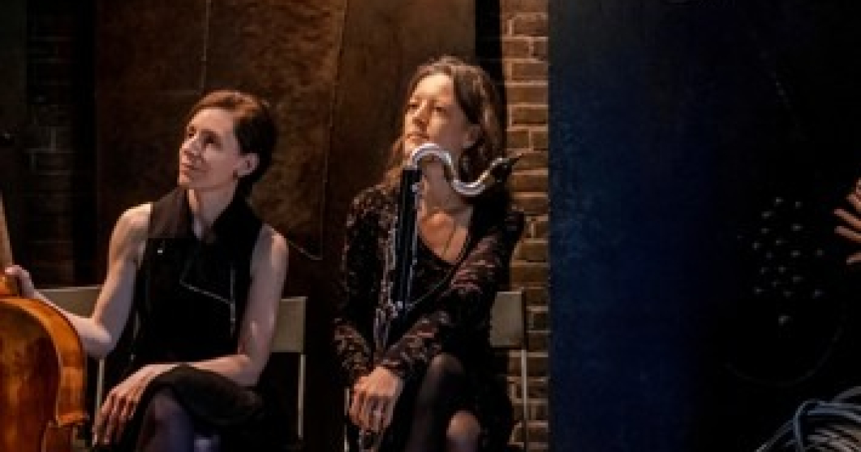 Katharina Gross en Fie Schouten zitten naast elkaar op het podium met hun instrumenten: de cello en de basklarinet. Ze dragen donkere kleding en kijken geamuseerd naar de linkerkant. Katharina Gross en Fie Schouten zitten naast elkaar op het podium met hun instrumenten: de cello en de basklarinet. Ze dragen donkere kleding en kijken geamuseerd naar de linkerkant.