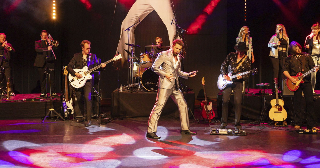 Bouke staat met de ElvisMatters Band op een rood verlicht podium in een gouden pak. Bouke staat met de ElvisMatters Band op een rood verlicht podium in een gouden pak.