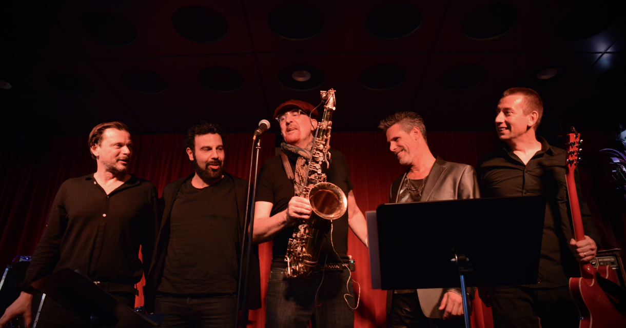 Bandfoto met de bandleden in donkere achtergrond met rood doek en een spot op hun gericht passend bij hun Bowie interpretatie.