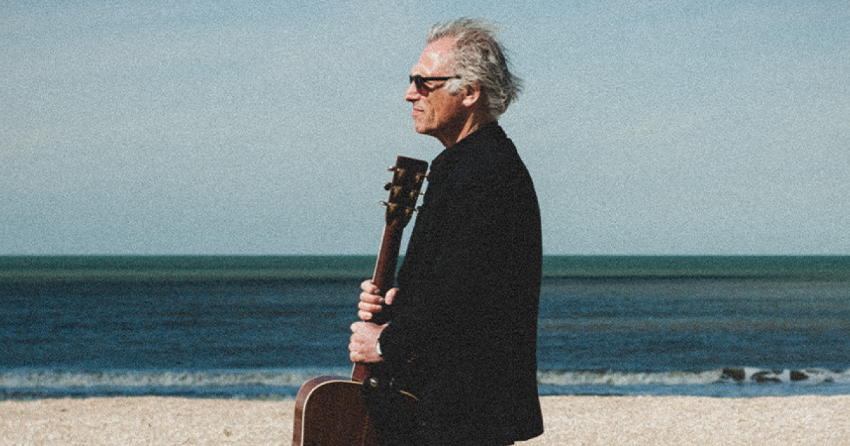 Philip Kroonenberg met gitaar in de hand op het strand, in de wind kijkend met zonnebril wat past bij emotie van zijn country genre.