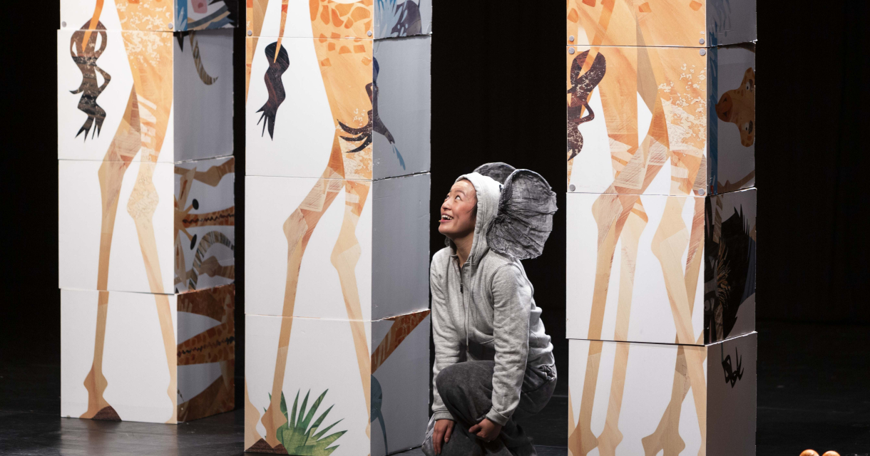 Een vrouw verkleed als olifant zit op een podium omringt door decorstukken die giraffen voorstellen. Een vrouw verkleed als olifant zit op een podium omringt door decorstukken die giraffen voorstellen.