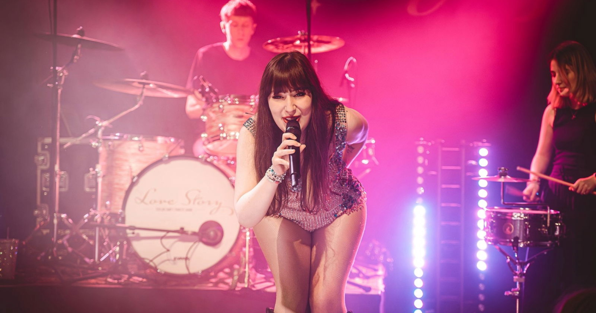 De leden van de Taylor Swift Tribute Band staan op een roze verlicht podium. De zangeres kijkt lachend de camera in. De leden van de Taylor Swift Tribute Band staan op een roze verlicht podium. De zangeres kijkt lachend de camera in.