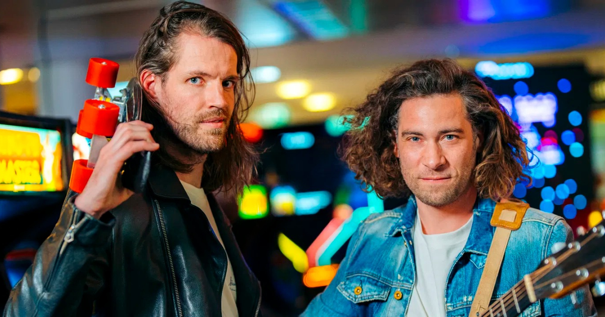 De leden van Popup choir, Maarten Bos en gitarist Tijmen de Koning, staan met serieuze blikken in een ruimte vol arcade machine's.  