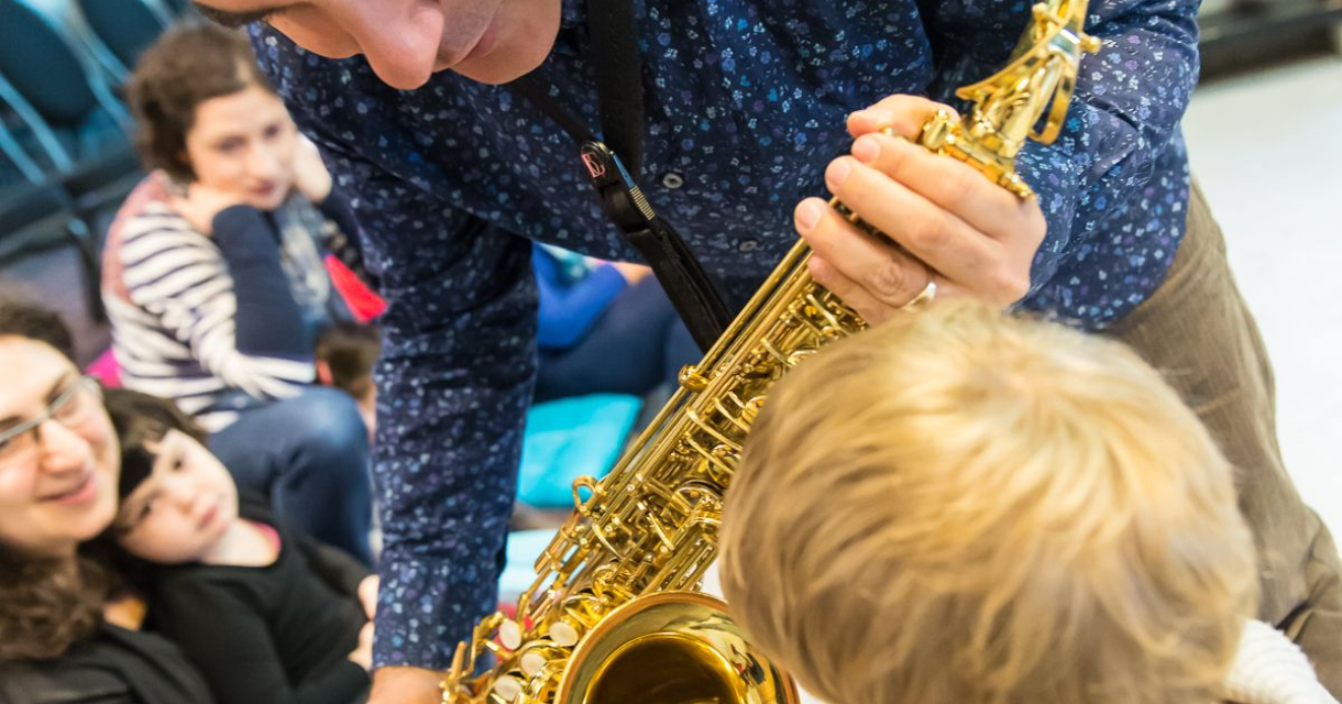 Een peuter kijkt naar de saxofoon van een muzikant tijdens het Ukkieconcert in Theater de Regentes Een peuter kijkt naar de saxofoon van een muzikant tijdens het Ukkieconcert in Theater de Regentes