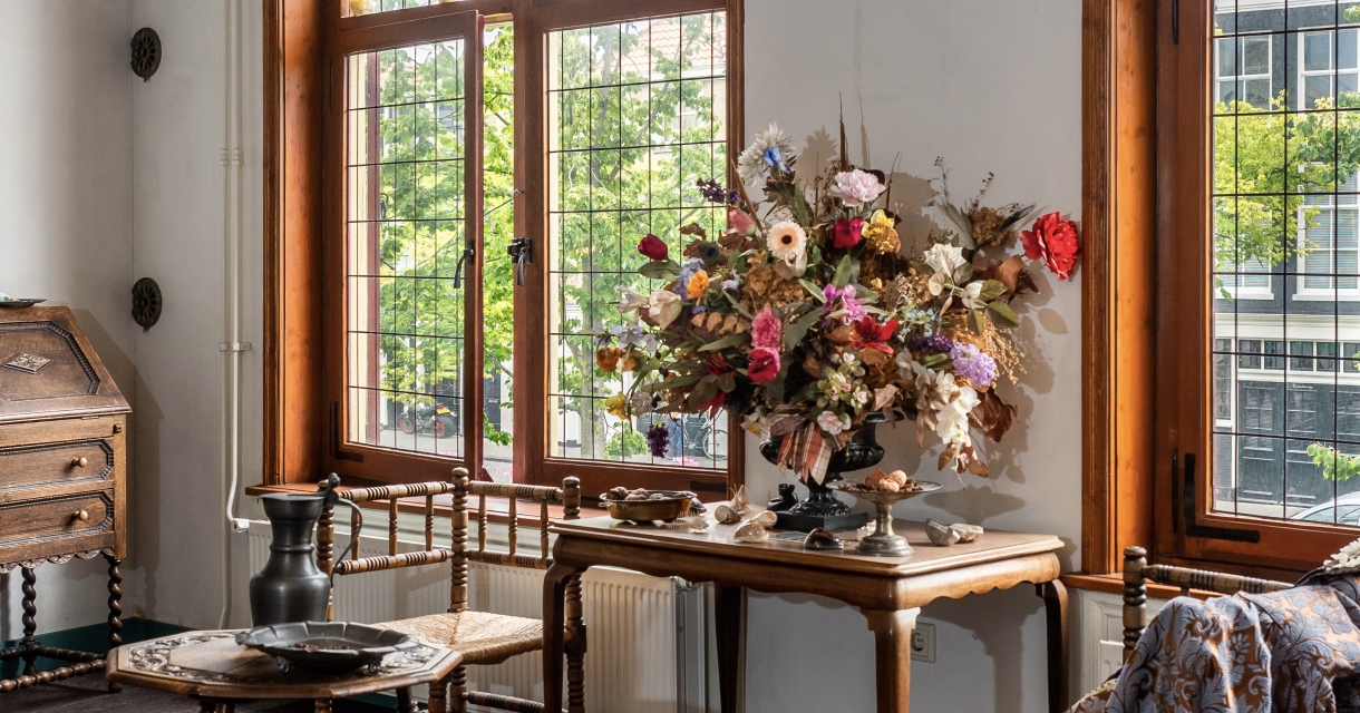 Historisch interieur met houten meubels, droogbloemen op een tafel en uitzicht op grachtenpand door glas-in-loodramen Historisch interieur met houten meubels, droogbloemen op een tafel en uitzicht op grachtenpand door glas-in-loodramen