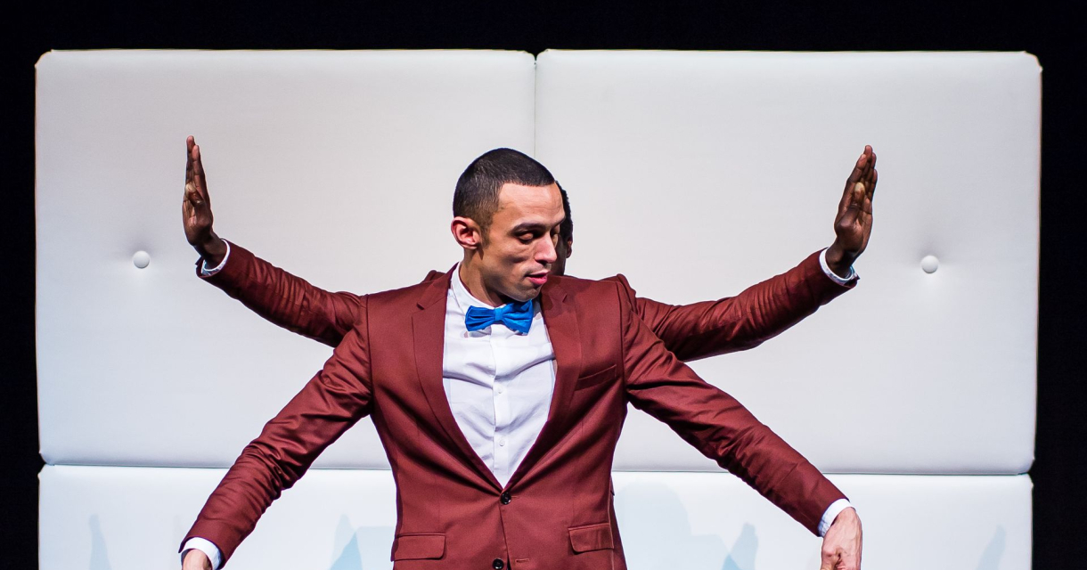 Man in rood pak met blauwe strik op podium, met extra armen zichtbaar door tweede persoon achter hem Man in rood pak met blauwe strik op podium, met extra armen zichtbaar door tweede persoon achter hem