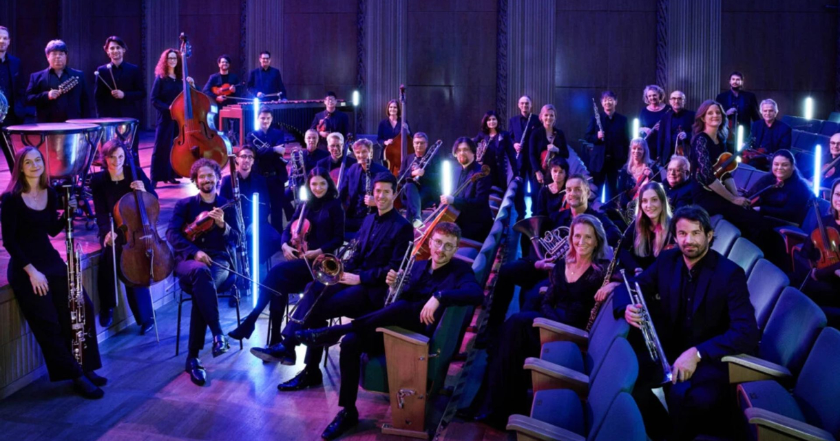 Leden van het WDR Funkhausorchester zitten met hun instrumenten op stoelen in een theaterzaal Leden van het WDR Funkhausorchester zitten met hun instrumenten op stoelen in een theaterzaal