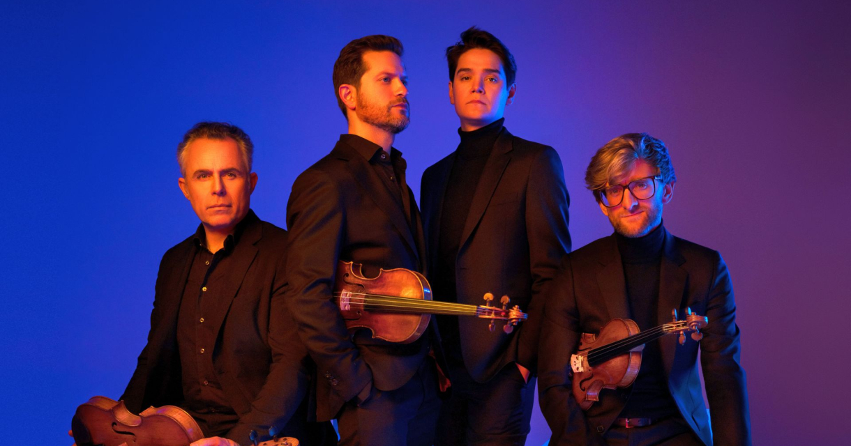 Vier mannen in zwarte pakken met strijkinstrumenten, poserend tegen een blauw-paars verlichte achtergrond Vier mannen in zwarte pakken met strijkinstrumenten, poserend tegen een blauw-paars verlichte achtergrond