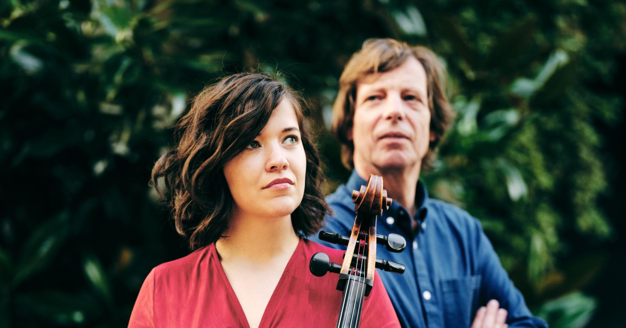Twee mensen staan buiten voor een groene achtergrond; de vrouw vooraan houdt een cello vast, de man staat erachter. Twee mensen staan buiten voor een groene achtergrond; de vrouw vooraan houdt een cello vast, de man staat erachter.