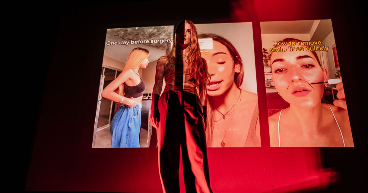 Een vrouw staat op een podium in rood licht, met projecties van drie verticale video’s van influencers op de achtergrond die schoonheidsproducten laten zien. Haar silhouet mengt met de beelden. Een vrouw staat op een podium in rood licht, met projecties van drie verticale video’s van influencers op de achtergrond die schoonheidsproducten laten zien. Haar silhouet mengt met de beelden.