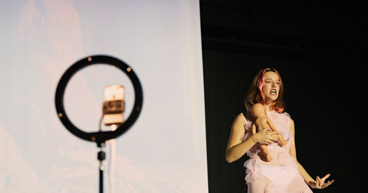 Vrouw in roze outfit performt voor een projectiescherm, met een ringlamp en smartphone op statief op de voorgrond. Vrouw in roze outfit performt voor een projectiescherm, met een ringlamp en smartphone op statief op de voorgrond.