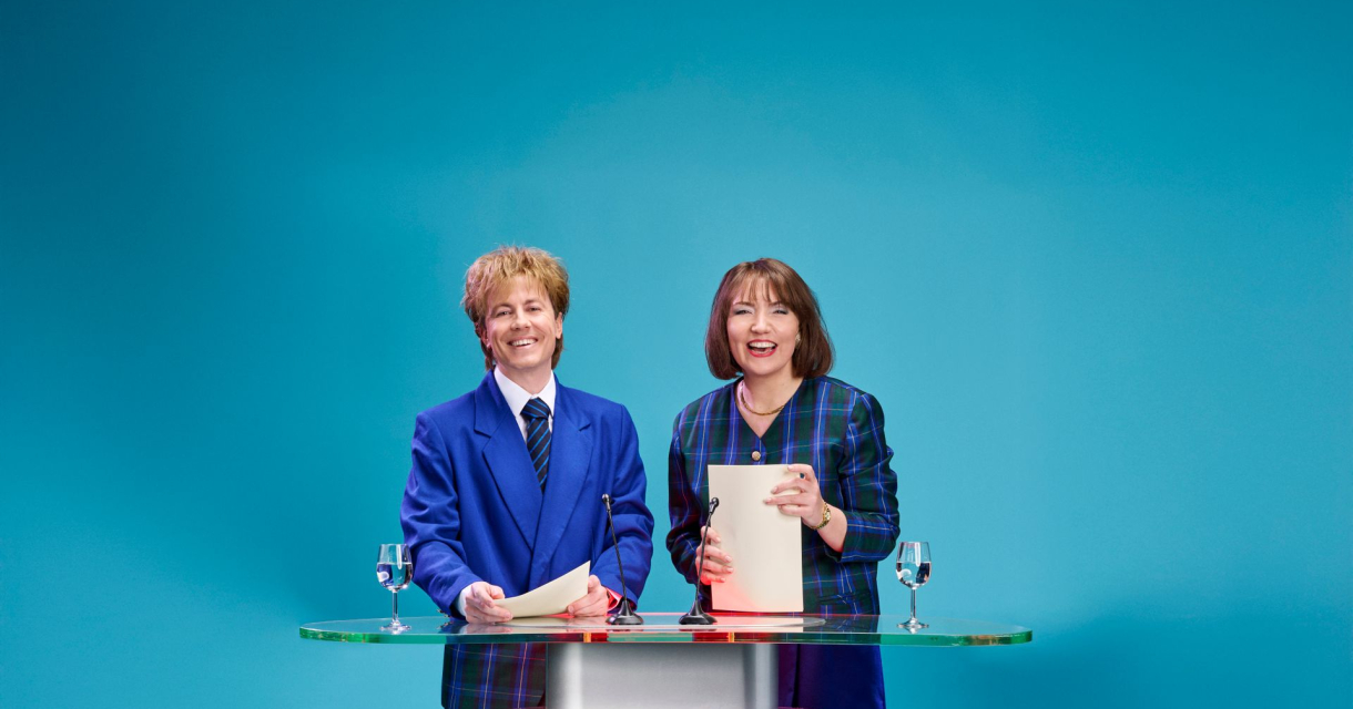 Twee nieuwslezers die aan een desk staan met een felblauwe achtergrond Twee nieuwslezers die aan een desk staan met een felblauwe achtergrond