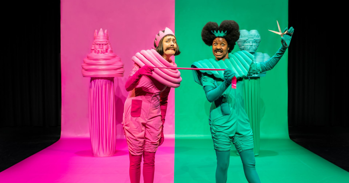 Twee acteurs in felroze en felgroen kostuum op gesplitst podium, met decor in bijpassende kleuren
