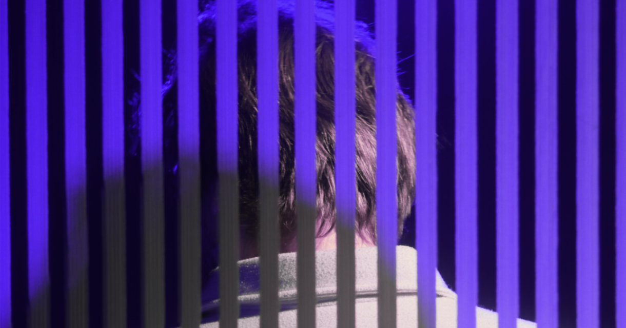 Achteraanzicht van een persoon in paars licht, gefilterd door verticale zwarte strepen die een gevangenis- of blindsuggestie oproepen. Achteraanzicht van een persoon in paars licht, gefilterd door verticale zwarte strepen die een gevangenis- of blindsuggestie oproepen.