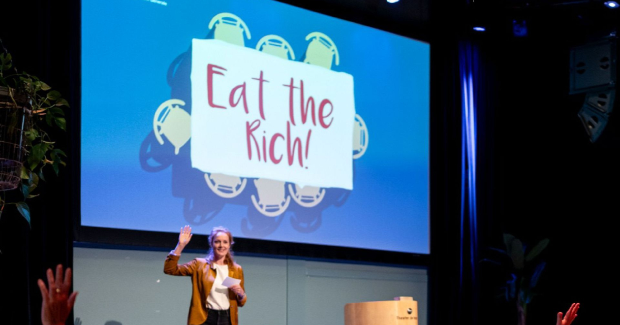 Spreker op podium voor groot scherm met tekst “Eat the Rich”, publiek steekt handen op in zaalsetting