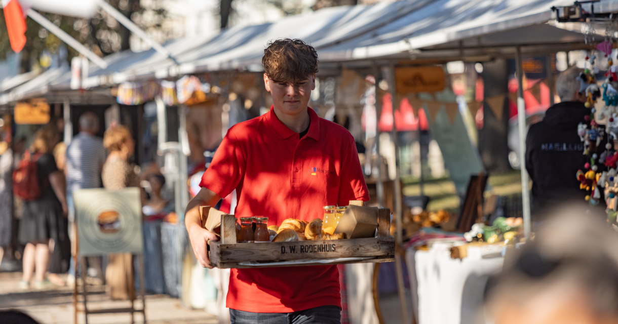 Een jongen in een rode polo draagt een dienblad met eten op een markt.