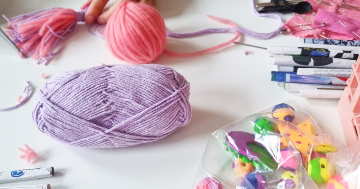 Een hand houdt een roze pompon en er ligt paars garen met knutselmateriaal op de tafel.