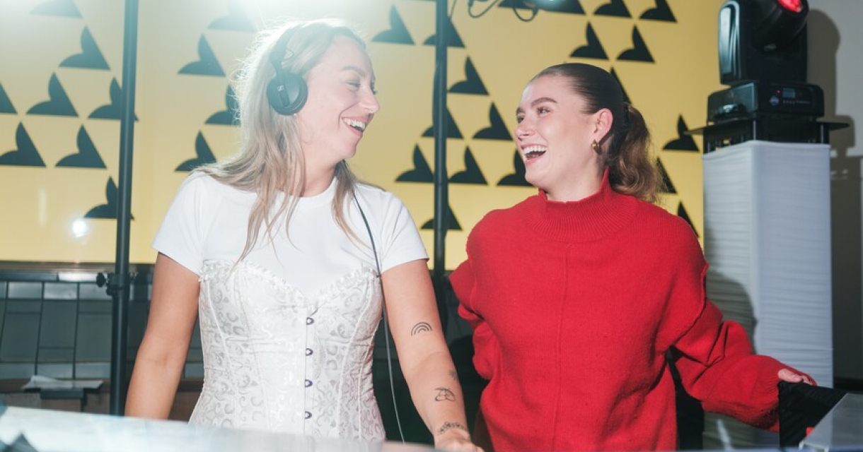 Twee lachende meisjes achter een dj tafel. Eén heeft een koptelefoon op. 
