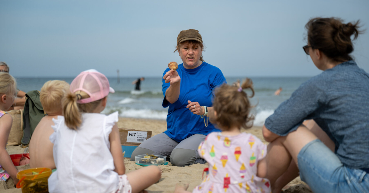 Een vrouw vertelt kinderen iets op het strand.