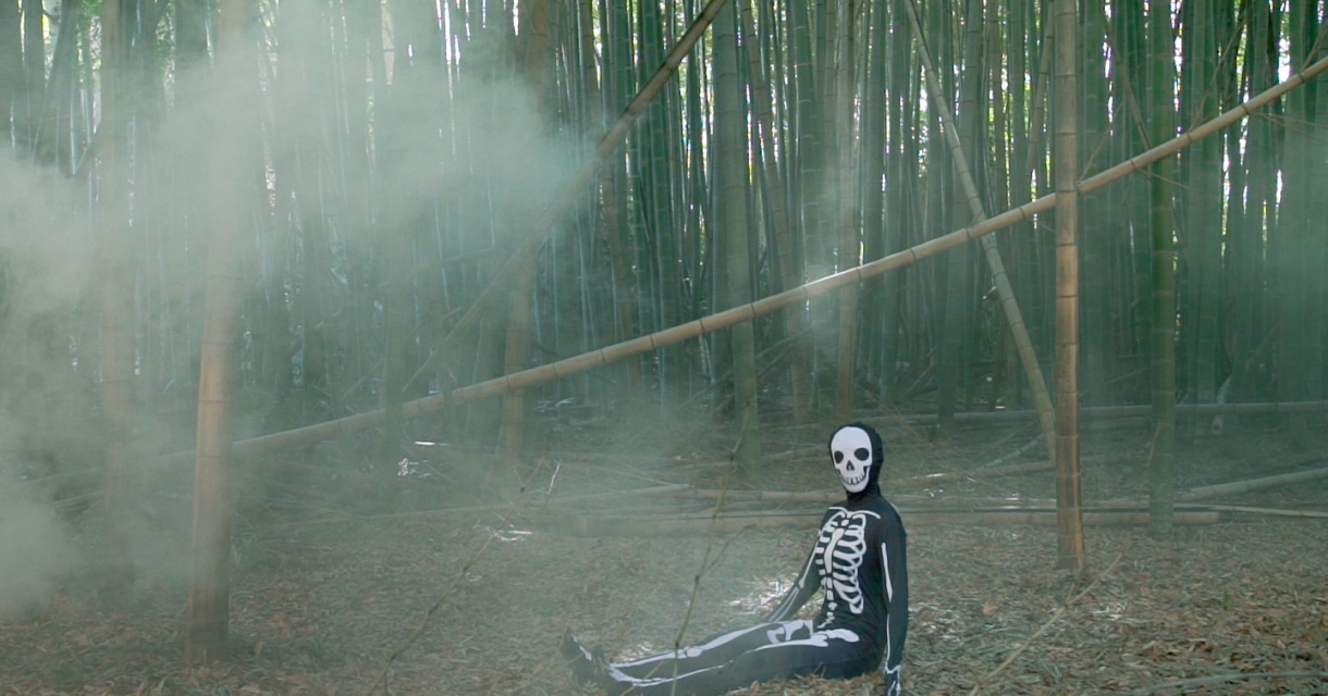 Een skelet in een zwart pak zit in een mistig bamboebos.