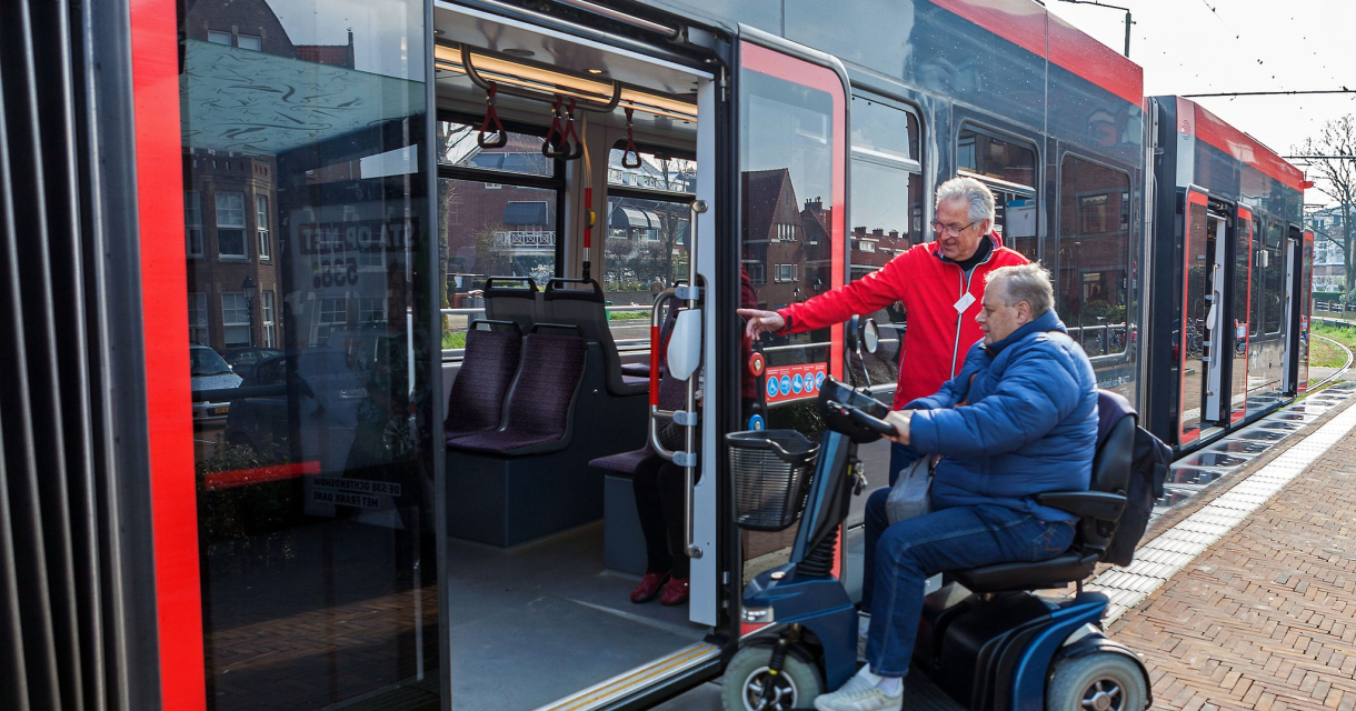 Een man in een scootmobiel stapt de tram binnen met hulp.