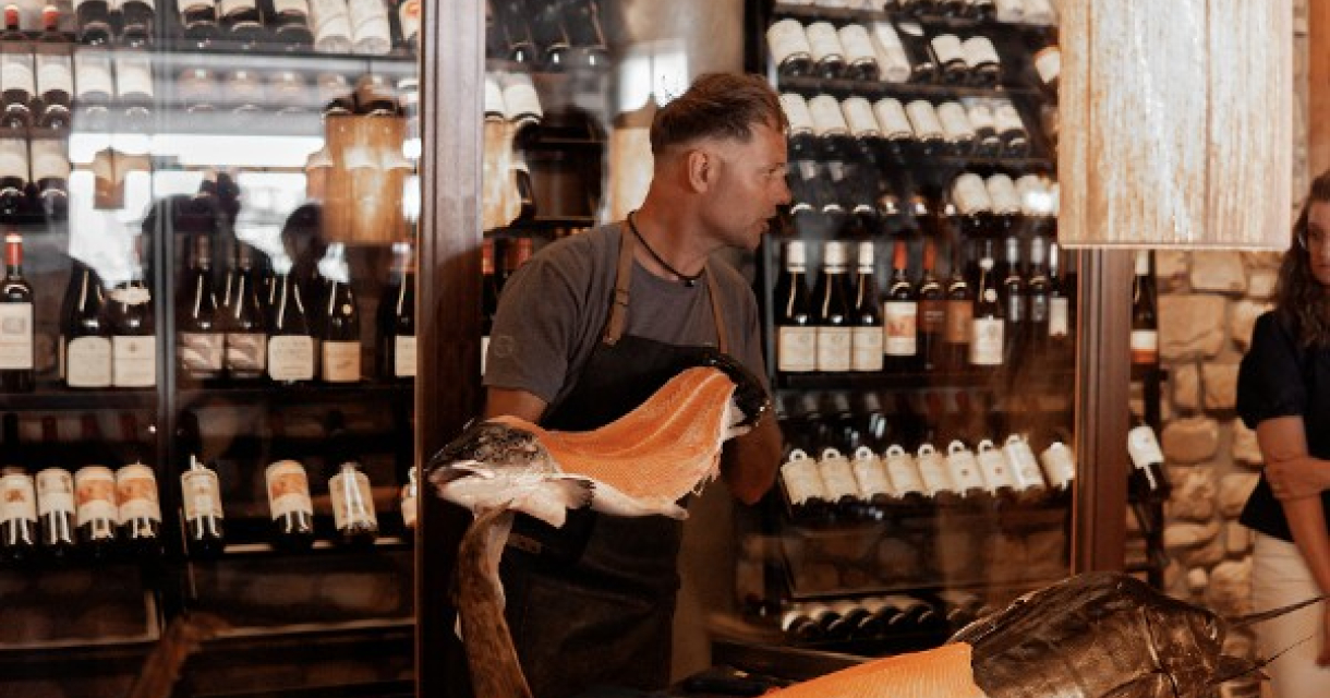 Een man staat in een restaurant met een gesneden vis en wijnflessen op de achtergrond.