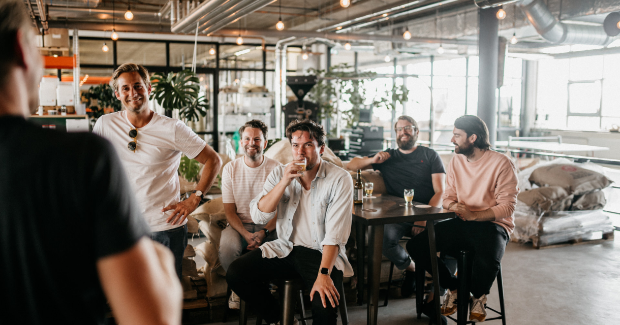 Vijf mannen zitten aan een tafel bij een bierproeverij. Ze lachen en drinken.
