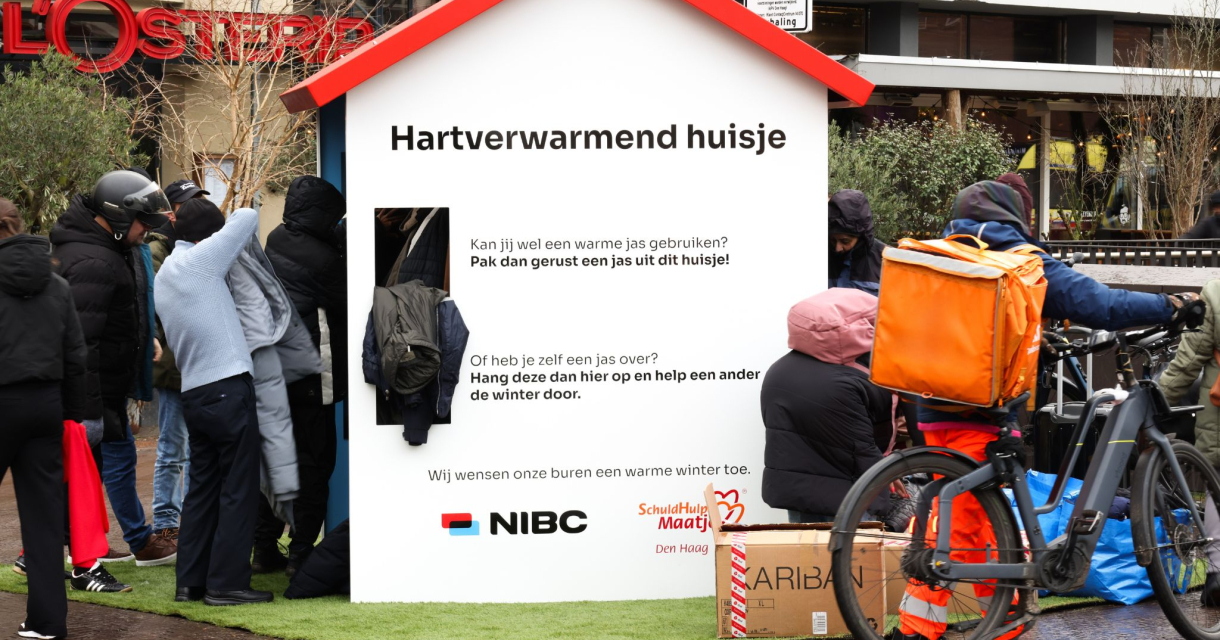 Houten huisje op de Grote Markt in Den Haag waar mensen omheen staan waaraan warme jassen worden uitgedeeld. 