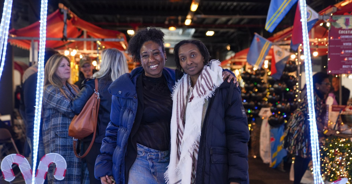 Twee vrouwen poseren samen op een kerstmarkt, omringd door kraampjes en feestelijke verlichting.