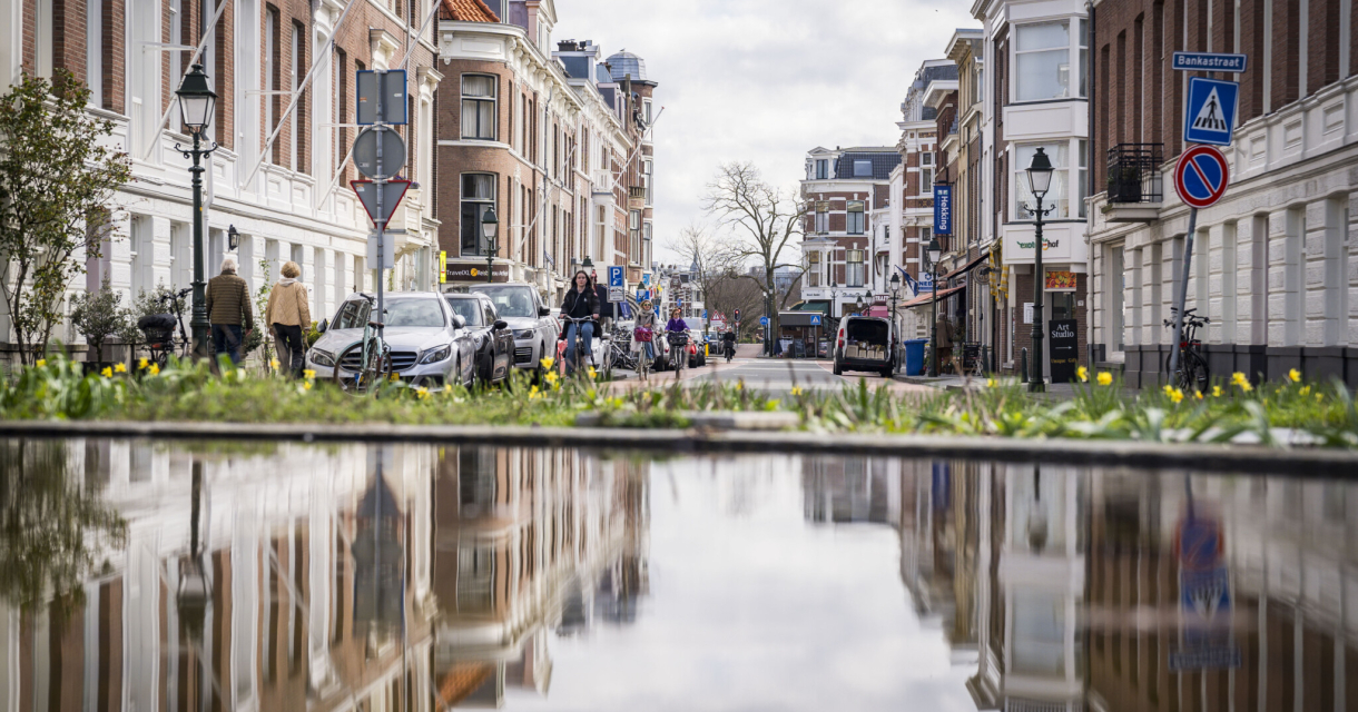 Een drukke straat in Den Haag met voetgangers en auto's, weerspiegeld in een plas water.