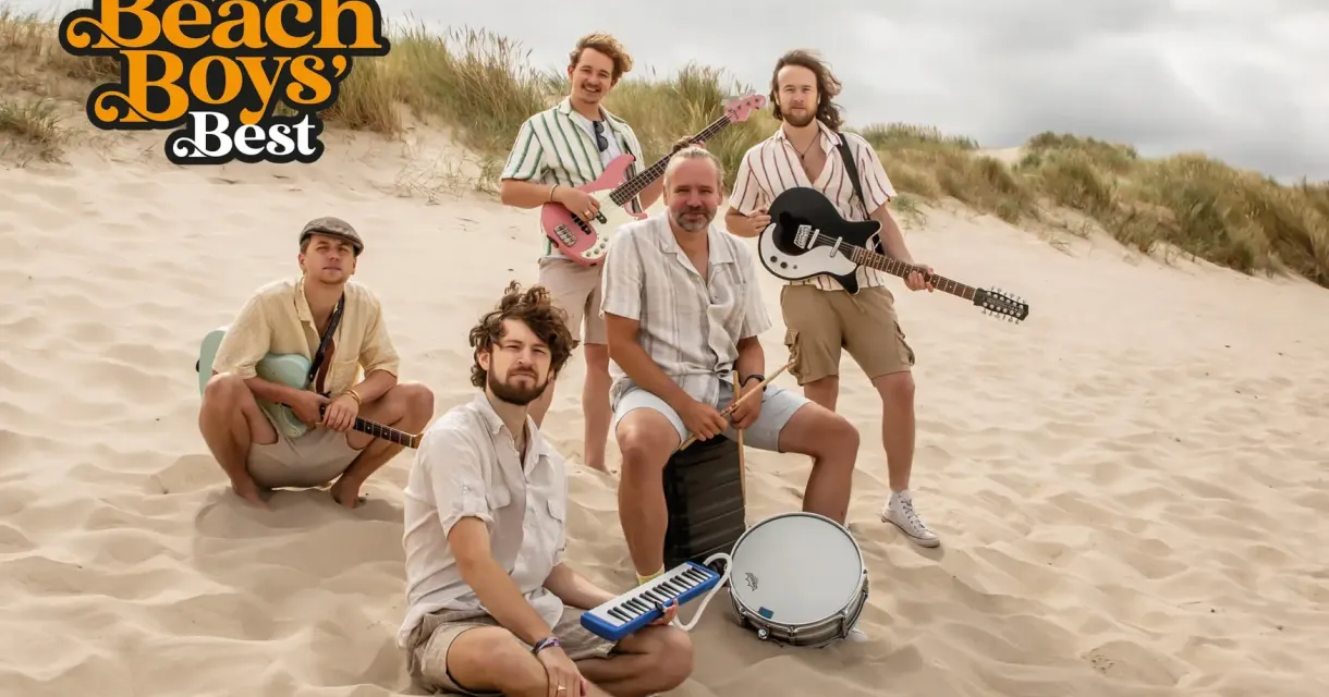 Vijf muzikanten zittend op een zandstrand met instrumenten.